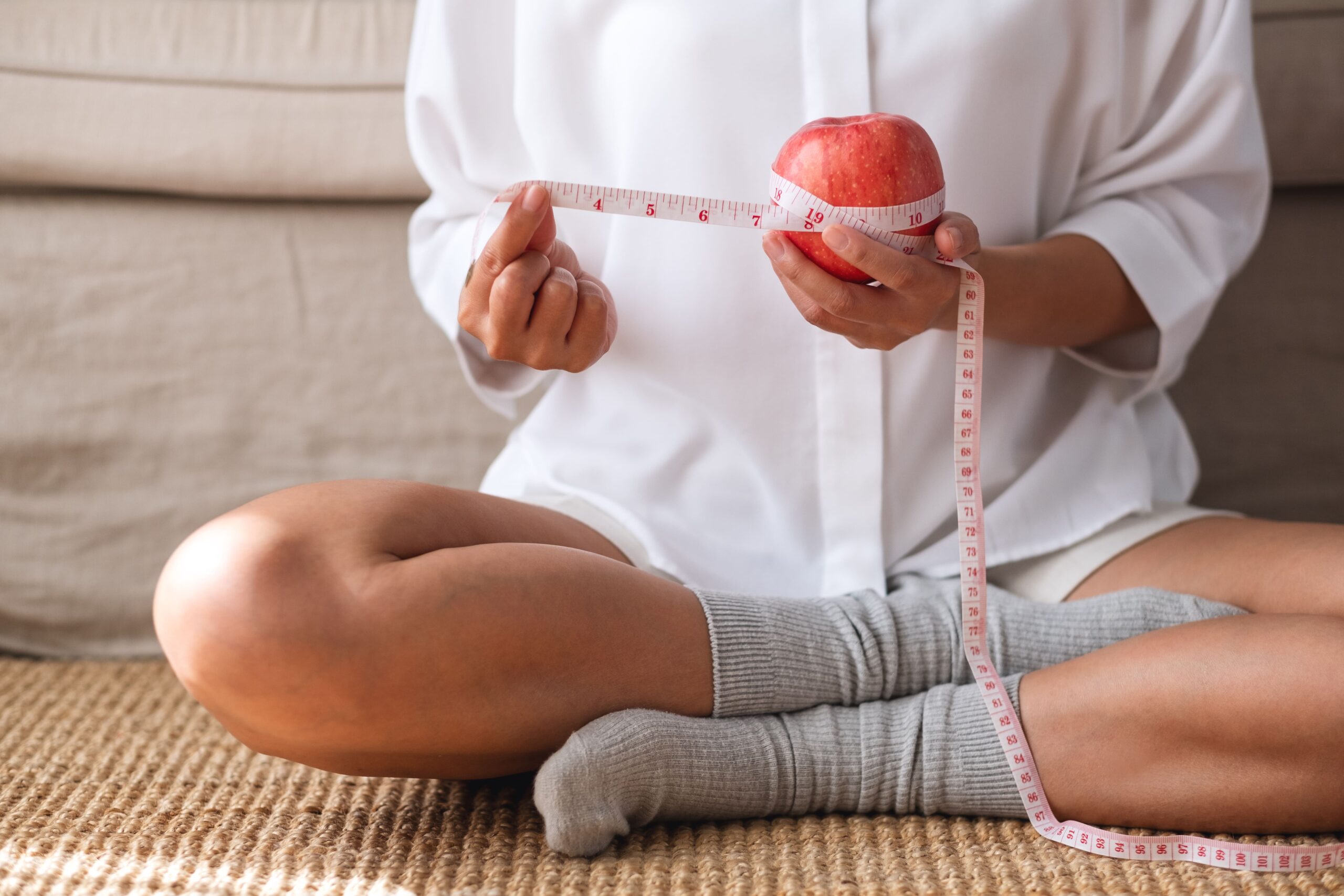 Close-up of a woman sitting cross-legged on a mat, holding an apple wrapped with a measuring tape, symbolizing nutrition, wellness, and healthy lifestyle choices.