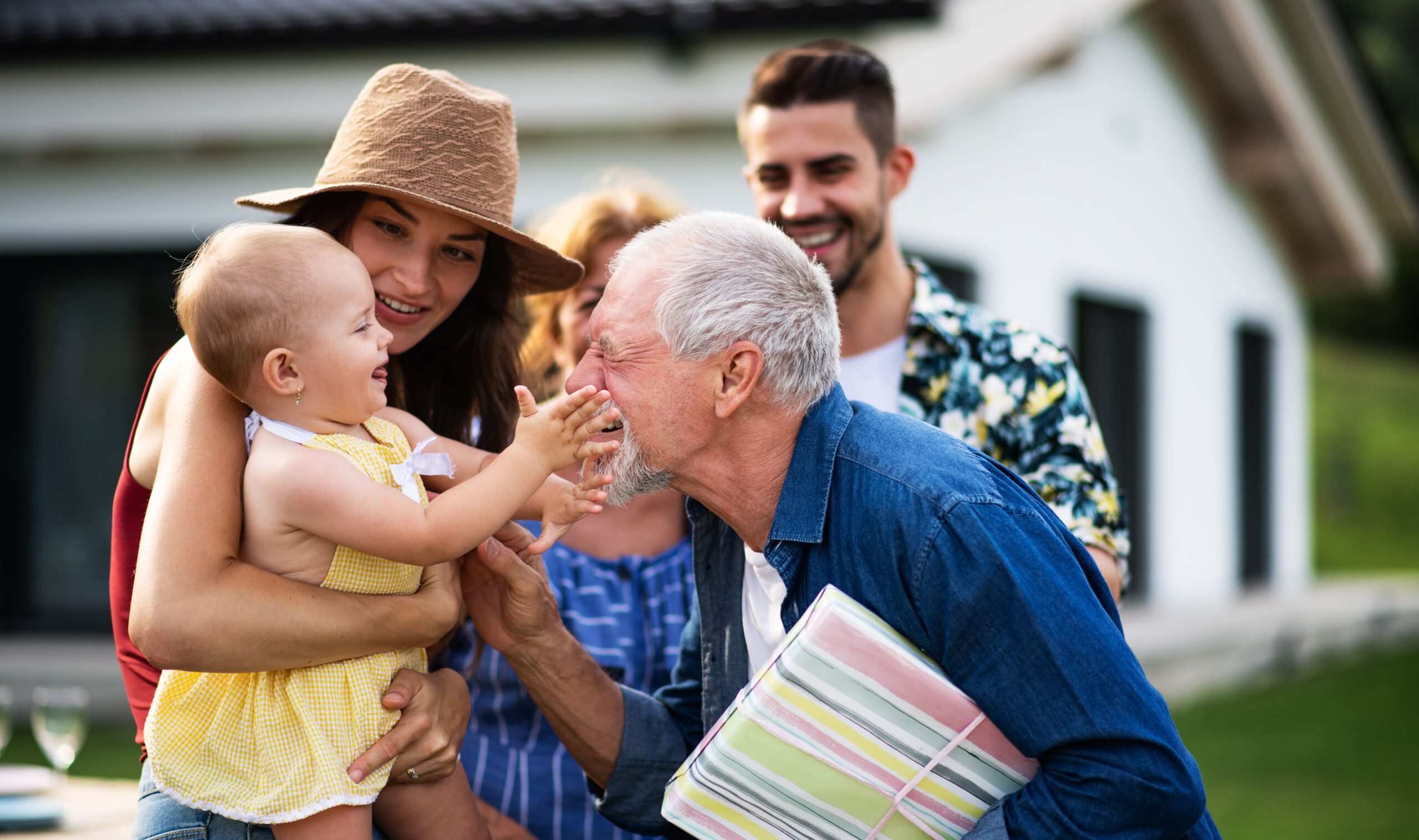 Multigenerational family outdoors, with a baby reaching for her grandfather’s face while parents and relatives smile in the background.