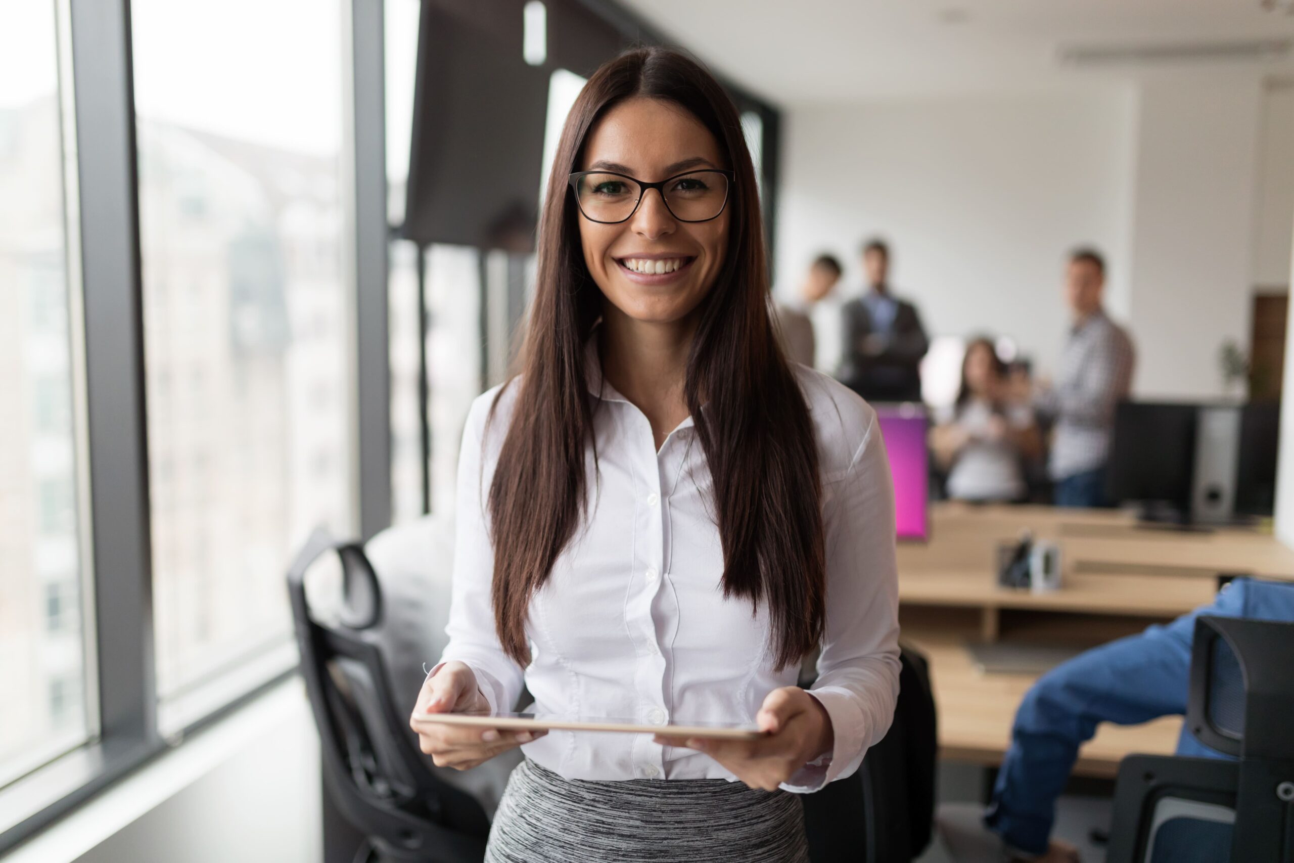 Confident HR professional standing in a bright, modern office holding a tablet and smiling, representing the role HR leaders play in building employee engagement and positive workplace culture.