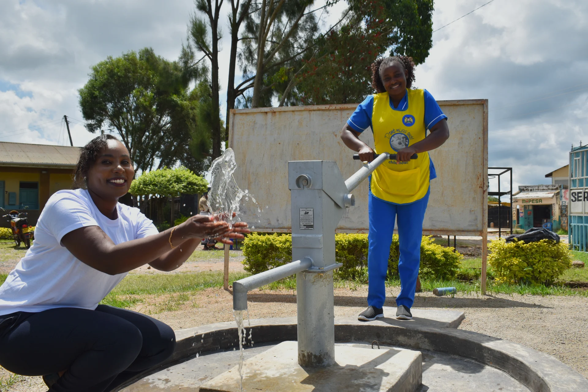 Two women in Kenya pump and collect clean water from a community well, smiling as they celebrate access to safe water provided through The Water Project.