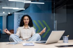 A woman in a modern office sits cross-legged at her desk, meditating with closed eyes and open hands, symbolizing workplace wellness and balance.