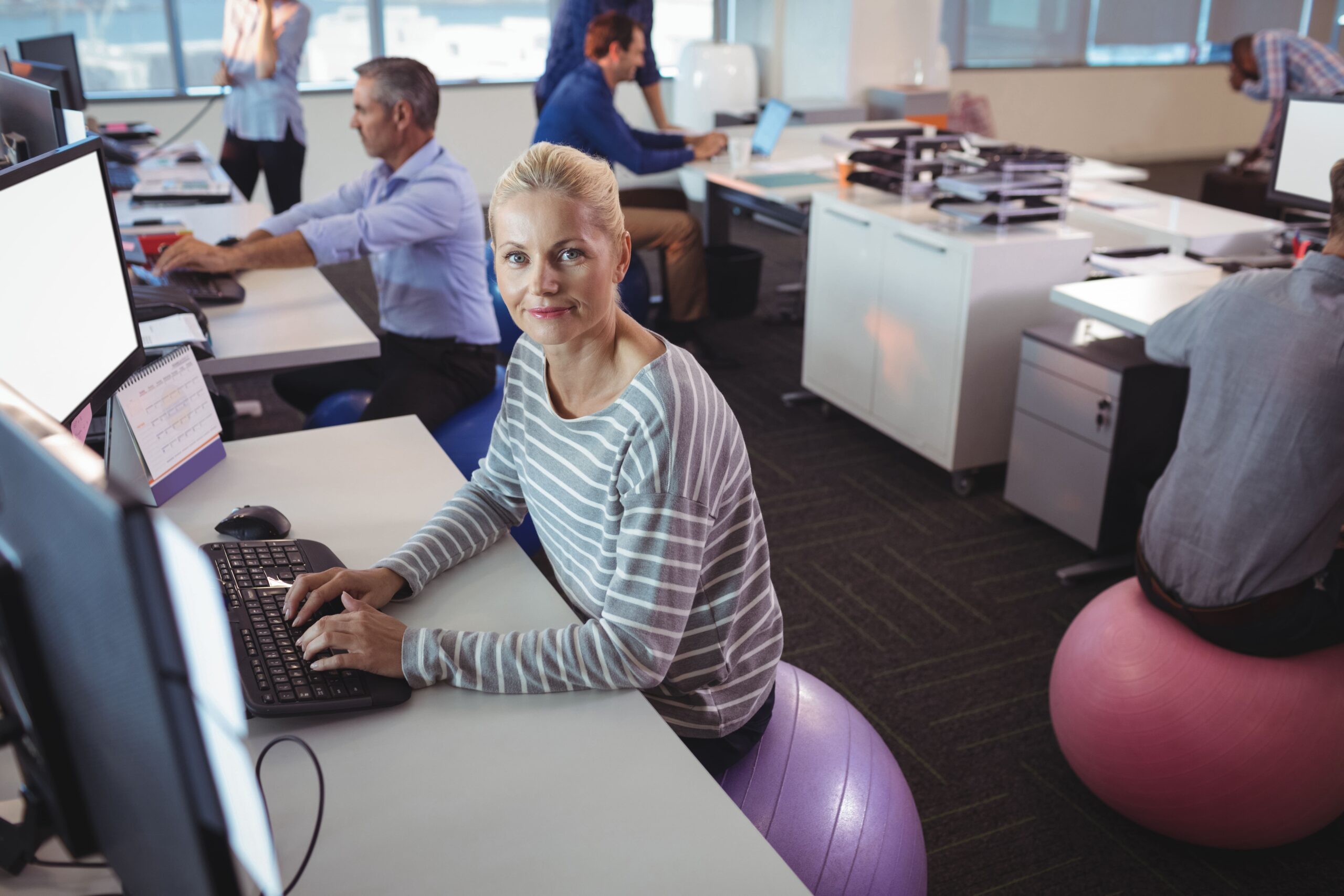 A smiling businesswoman works at her desk while sitting on an exercise ball in a modern office, promoting workplace wellness and active sitting habits.