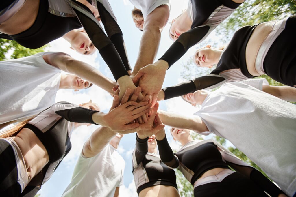 A team standing in a circle outdoors with their hands stacked together, symbolizing unity, teamwork, and the power of physical team-building activities.