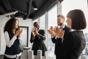 A diverse group of colleagues in business attire applauds together in a modern office, reflecting a culture of appreciation and teamwork.