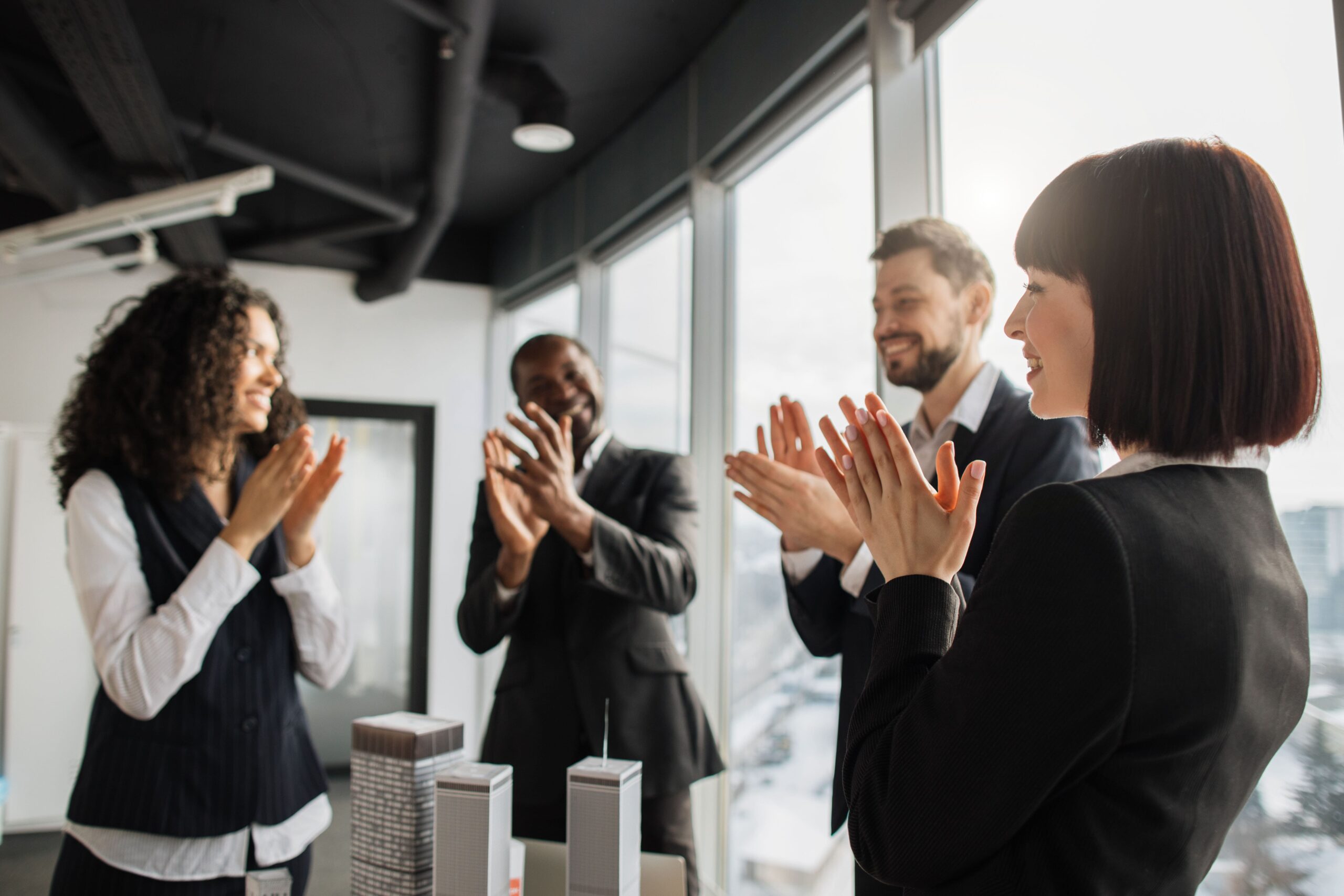 A diverse group of colleagues in business attire applauds together in a modern office, reflecting a culture of appreciation and teamwork.