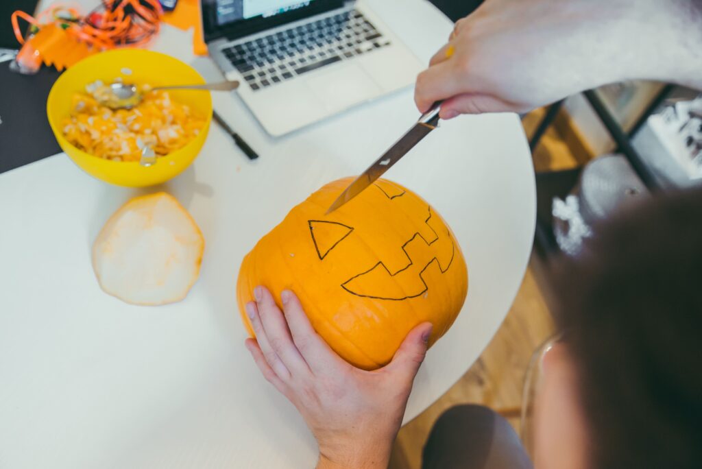 A person carves a jack-o’-lantern pumpkin on a desk with a laptop and bowl of pumpkin seeds nearby during a Halloween office activity.