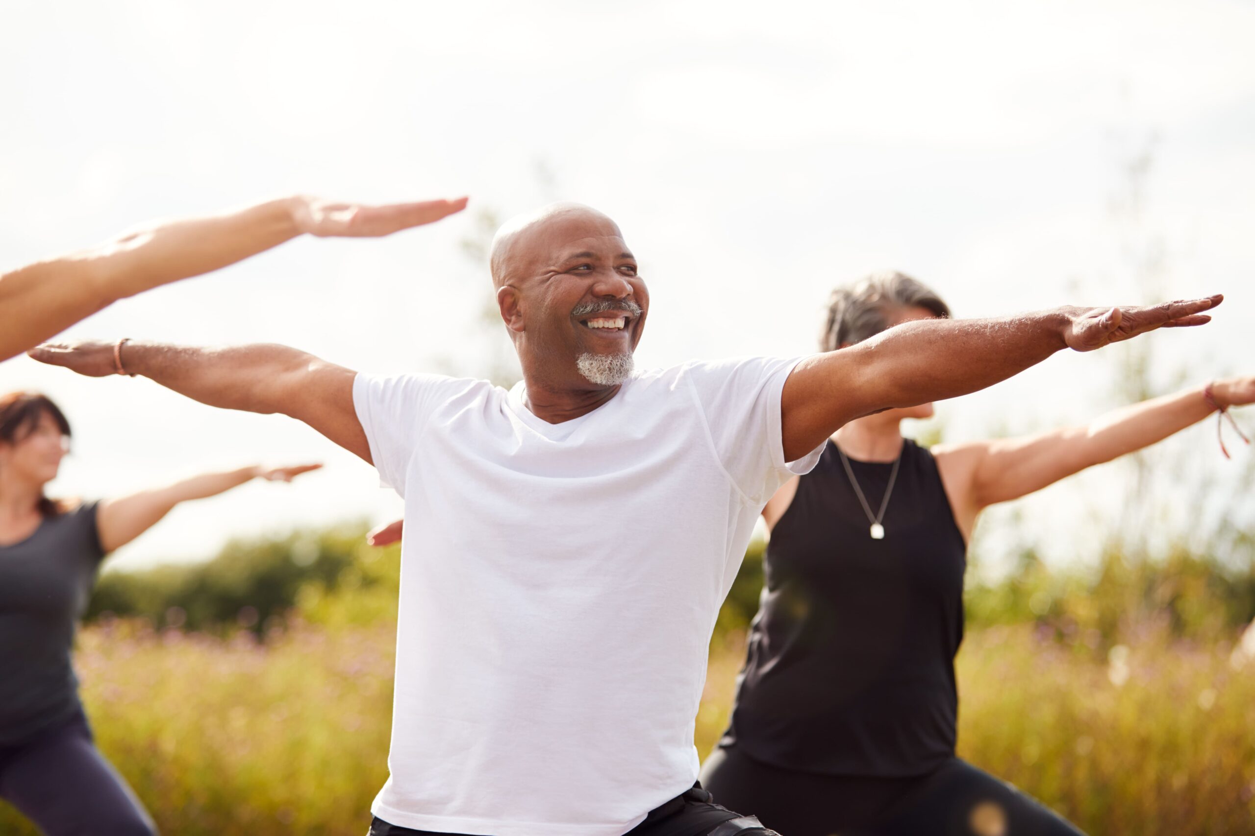 A group of adults participating in an outdoor yoga class, with a smiling man in the foreground practicing a stretching pose, representing wellness, movement, and preventative health programs for employers and TPAs.