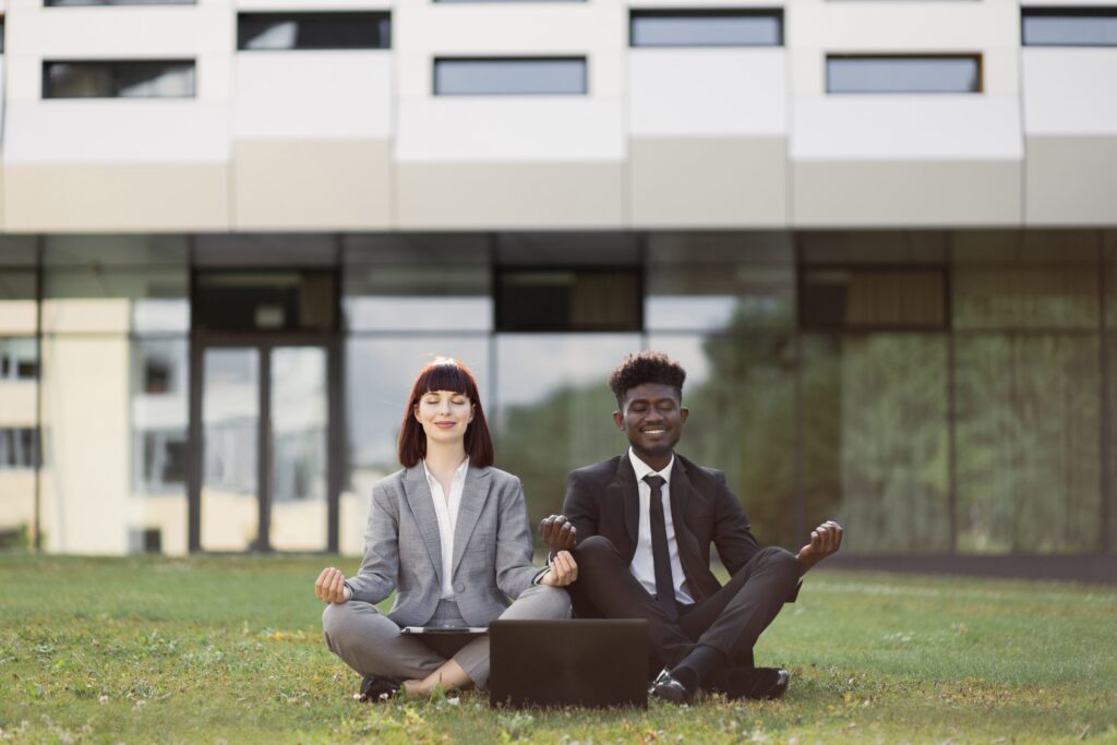 Two business professionals sit outdoors in a meditation pose, eyes closed and smiling, demonstrating workplace wellbeing and stress-reduction practices that support TPA employer retention strategies.