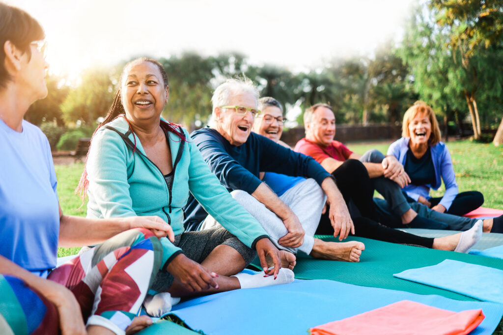 A diverse group of adults sitting on yoga mats outdoors, smiling and talking after a group wellness session, representing personalized wellbeing and community-focused health support.