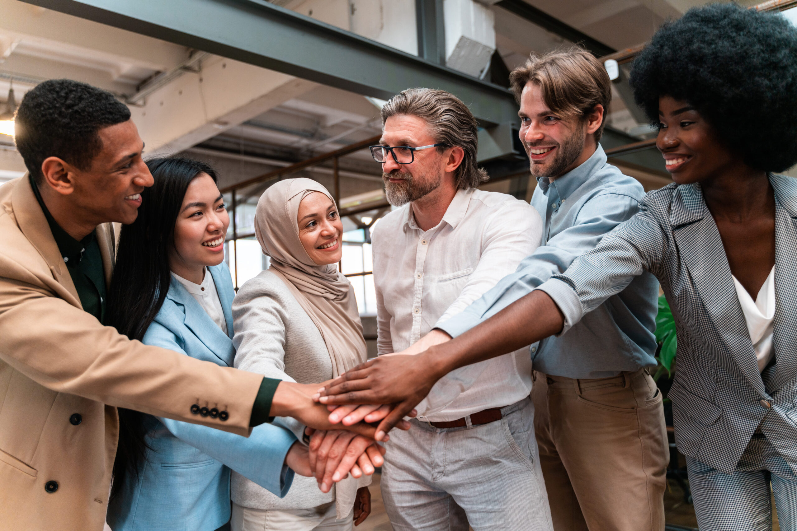 Diverse group of employees standing in a modern office with hands stacked together in the center, symbolizing teamwork, belonging, and a welcoming employee onboarding experience.