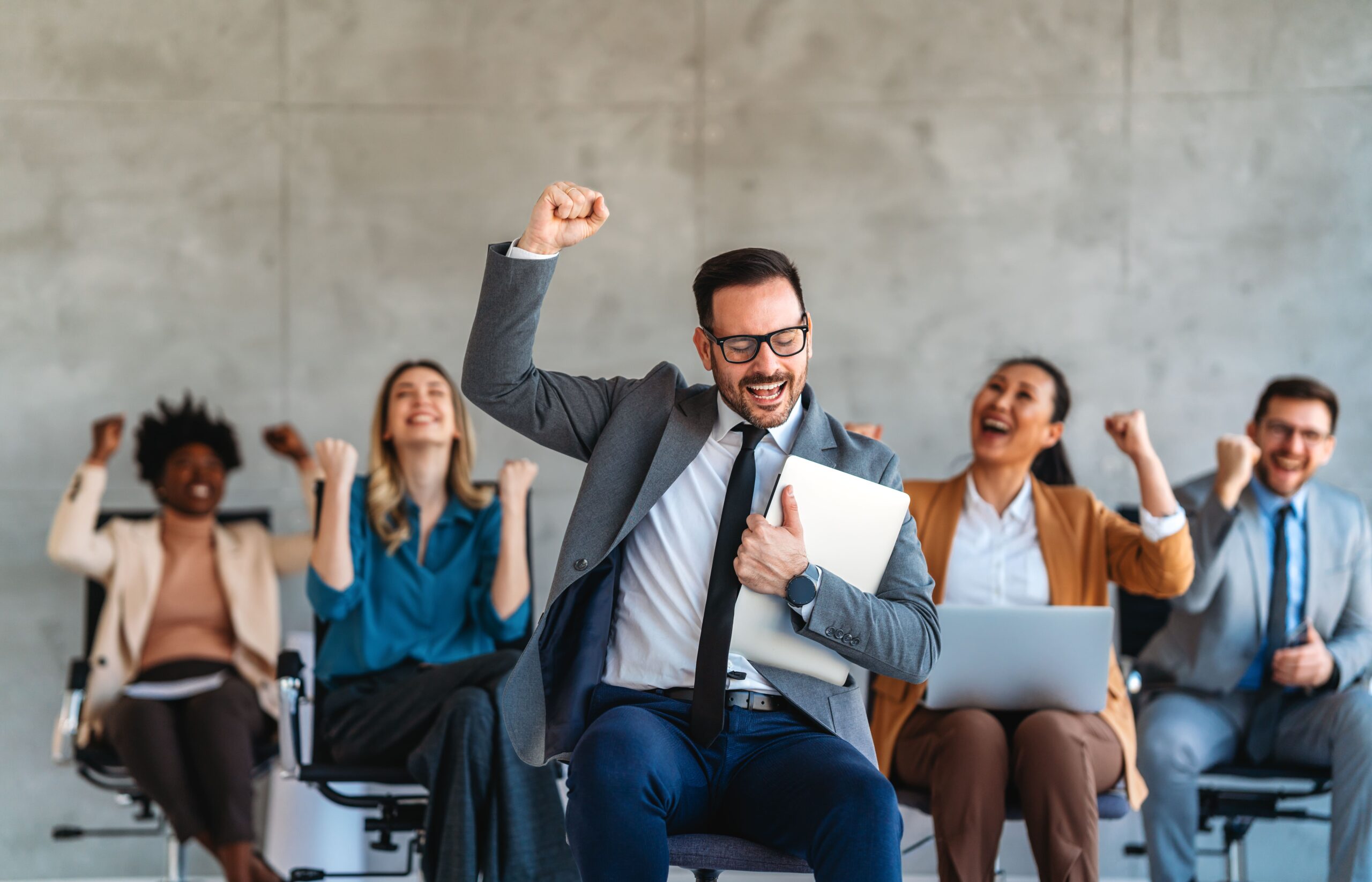 Smiling business professional celebrating success while colleagues cheer in the background, representing a modern employee experience built on engagement, motivation, and support.