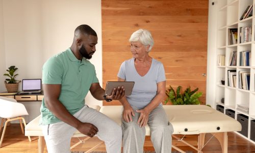 Physical therapist showing a personalized wellness plan on a digital tablet to an older woman during a consultation in a bright, modern clinic.