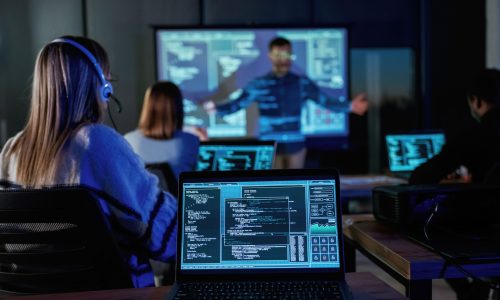 Employees participate in a cybersecurity training session, with laptops showing code and a presenter explaining threats on a projected screen.
