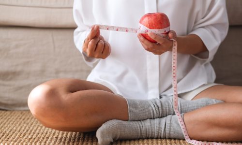 Close-up of a woman sitting cross-legged on a mat, holding an apple wrapped with a measuring tape, symbolizing nutrition, wellness, and healthy lifestyle choices.