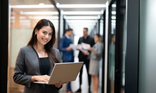 A smiling business professional stands in a modern office hallway working on a laptop, with colleagues meeting in the background—representing confident, competitive TPAs navigating industry changes.