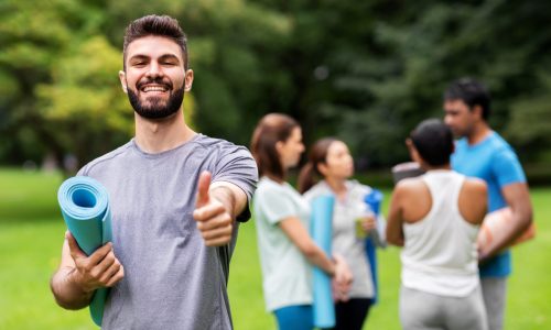 Smiling man holding a yoga mat and giving a thumbs-up in a park with a group of coworkers preparing for a wellness activity in the background, representing employee wellness and teamwork.