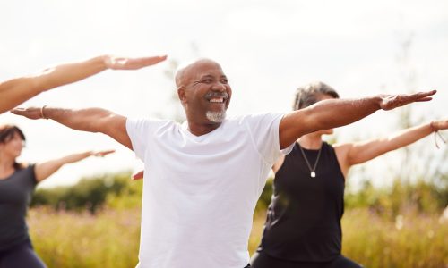 A group of adults participating in an outdoor yoga class, with a smiling man in the foreground practicing a stretching pose, representing wellness, movement, and preventative health programs for employers and TPAs.