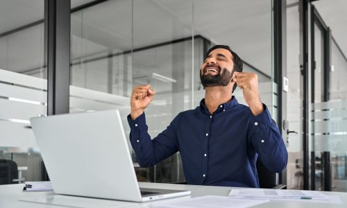 Smiling employee celebrating success while working on a laptop in a modern office, representing motivation and engagement from virtual employee incentives.