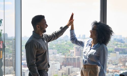 Two business professionals smiling and giving a high-five in a bright office with a city skyline in the background, symbolizing successful collaboration and achieving meaningful outcomes.