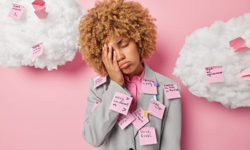 A stressed young woman in a suit stands against a pink background with her hand on her face, looking overwhelmed, covered in sticky notes listing tasks, symbolizing burnout and overload.