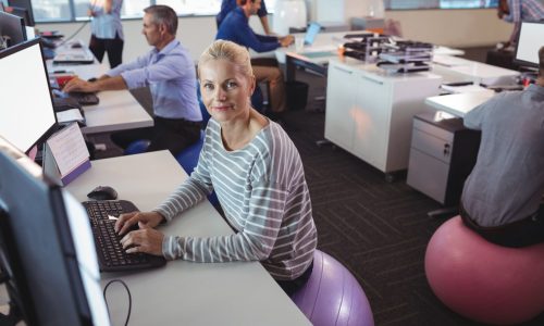 A smiling businesswoman works at her desk while sitting on an exercise ball in a modern office, promoting workplace wellness and active sitting habits.