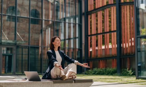 A professional woman sits outdoors in a meditation pose with her laptop beside her, symbolizing wellbeing and prevention as essential components of modern TPA value-added services.