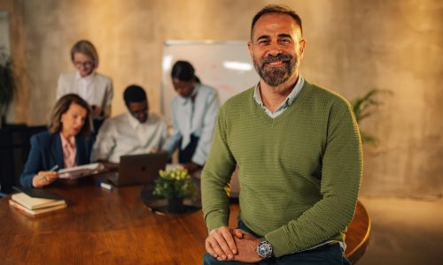 Confident business leader smiling while seated at a conference table, with a diverse team collaborating in the background — representing leadership resilience and team wellbeing in a startup environment.