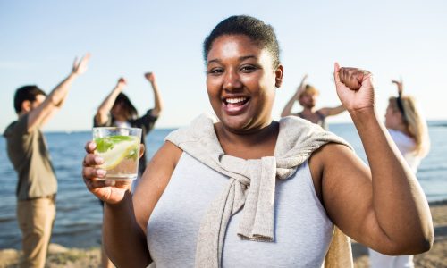 A smiling woman enjoys a drink at a beach gathering while friends celebrate in the background, symbolizing wellbeing and healthier lifestyles that help reduce diabetes-related claims.