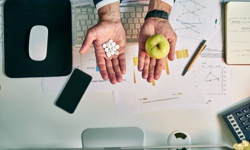A business professional sits at a desk holding white medication pills in one hand and a green apple in the other, symbolizing the choice between medical treatment and preventive wellness amid healthcare data and reports.