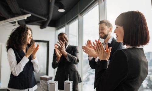 A diverse group of colleagues in business attire applauds together in a modern office, reflecting a culture of appreciation and teamwork.