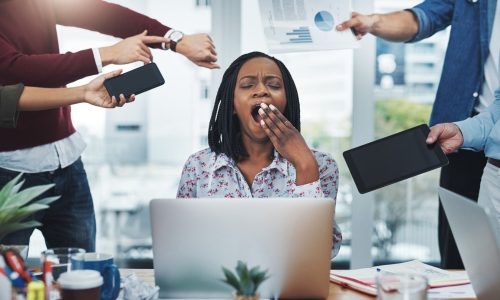 Stressed startup founder yawning at her desk surrounded by coworkers demanding attention with charts, devices, and time pressure — symbolizing burnout and the toll of constant demands.