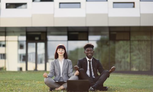 Two business professionals sit outdoors in a meditation pose, eyes closed and smiling, demonstrating workplace wellbeing and stress-reduction practices that support TPA employer retention strategies.