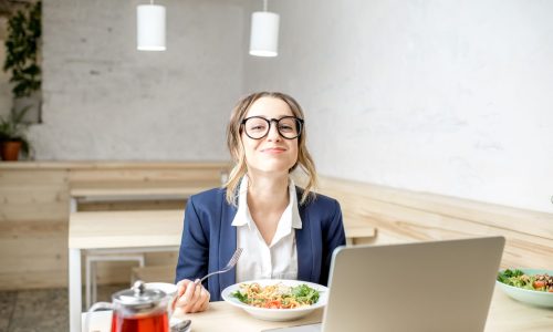 Smiling businesswoman enjoying a healthy lunch at her desk while working on a laptop in a bright office café.