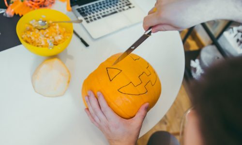 A person carves a jack-o’-lantern pumpkin on a desk with a laptop and bowl of pumpkin seeds nearby during a Halloween office activity.